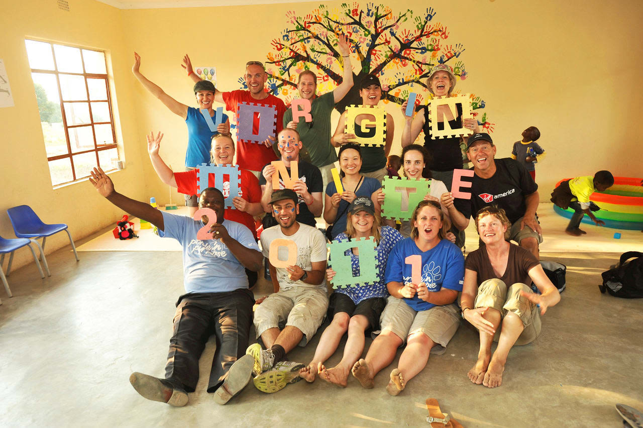 A group of people stand and sit in front of a tree mural on a yellow wall, each holding a letter or number to spell out 'Virgin Unite 2011'.
