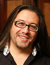 Color photograph of the face of a smiling man with long black hair and glasses