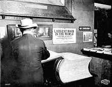 An extremely large book rests on a table, with its front cover and a small portion of its pages flipped open to the left. A man facing away from the camera stands in front of the flipped part. On the wall above the book is a sign saying "LARGEST BOOK IN THE WORLD - VISITORS' REGISTER FOR CALIFORNIA BUILDING - Alaska–Yukon–Pacific Exposition […]". On a table in the background and to the right of the large book is another table on which many small stacks of normal-sized books are visible.