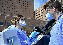 Two medical professionals, one holding a clipboard, in blue scrubs and facemasks stand outside the window of a dark blue car parked in front of a brick building.