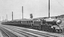 A black and white picture of four railway lines in a shallow cutting, a large steam engine leads a train of coaches from middle-left to right-foreground