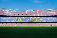 The words "Més que un club" are painted in yellow on the blue seats of the stadium