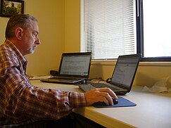 A grey-haired person using a computer with two monitors.