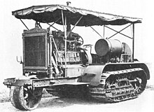 An early bulldozer-like tractor, on crawler tracks, with a leading single wheel – for steering – projecting from the front on an extension to the frame. The large internal combustion engine is in full view, with the cooling radiator prominent at the front. An overall roof is supported by thin rods, and side protection sheeting is rolled up under the edge of the roof.