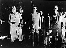 A group of actors in zombie makeup shamble across the unlit lawn of the farmhouse.