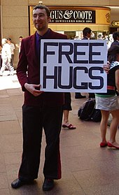 Color photograph of a man holding a large sign saying "FREE HUGS" in a mall