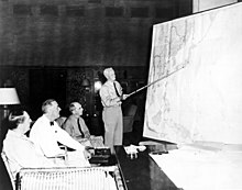 Three men are seated in lounge chairs. One is standing, holding a long stick and pointing to the location of Japan on a wall map of the Pacific.