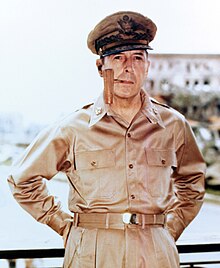 MacArthur in khaki trousers and open necked shirt with five-star-rank badges on the collar. He is wearing his field marshal's cap and smoking a corncob pipe.