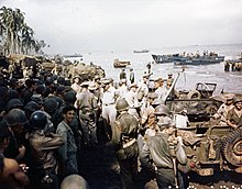 A large crowd of soldiers and jeeps on a beach. There are palm trees in the distance and landing craft offshore. A small group in the center conspicuously wear khaki uniforms and peaked caps instead of jungle green uniforms and helmets.