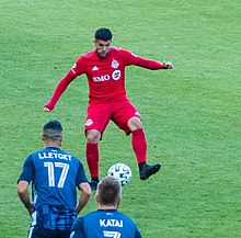 Alejandro Pozuelo, wearing a red Toronto FC jersey, dribbles a ball betweeb his feet in front of two opposing players