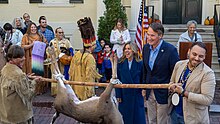 An older white man in a dark blue blazer smiles as he is presented with a dead deer hanging upside down held by two men in contemporary Native American atire.