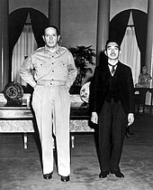 A tall Caucasian male (MacArthur), without hat and wearing open-necked shirt and trousers, standing beside a much shorter Asian man (Hirohito) in a dark suit.