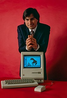 With a red background, Steve Jobs rests his forearms on a Macintosh computer.
