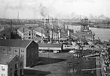 Several World War I ships line a port crowded with warehouses, with a city skyline behind them.
