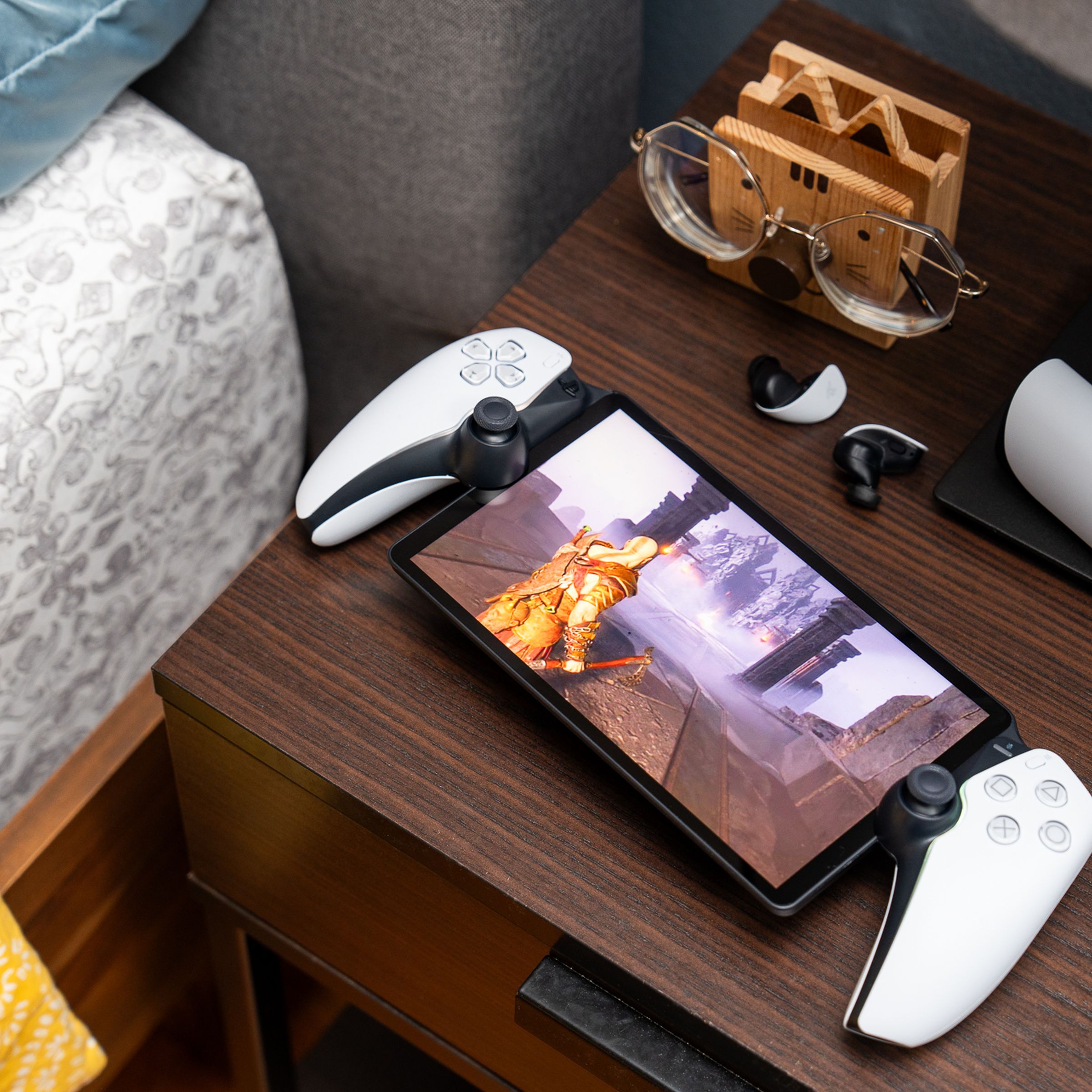 The PlayStation Portal sitting on a bedside table with a pair of earbuds. The handheld gaming device is streaming God of War: Ragnarök off a PlayStation 5.