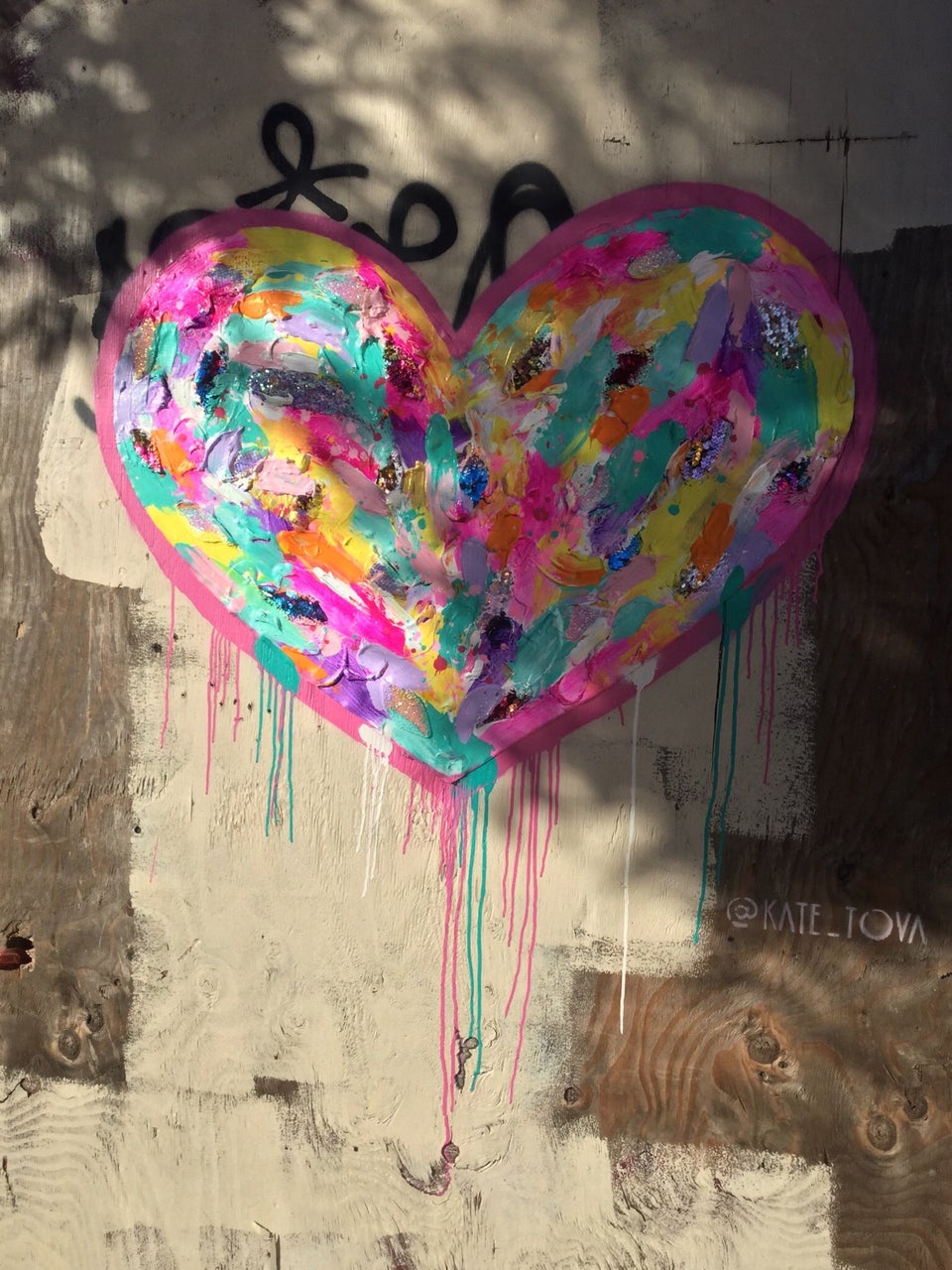Multicolored heart painting with a few sequine pieces, outlined in pink with bits of turquoise, paint dripping downward, on a piece of plywood at a construction site, signed KATE_TOVA.