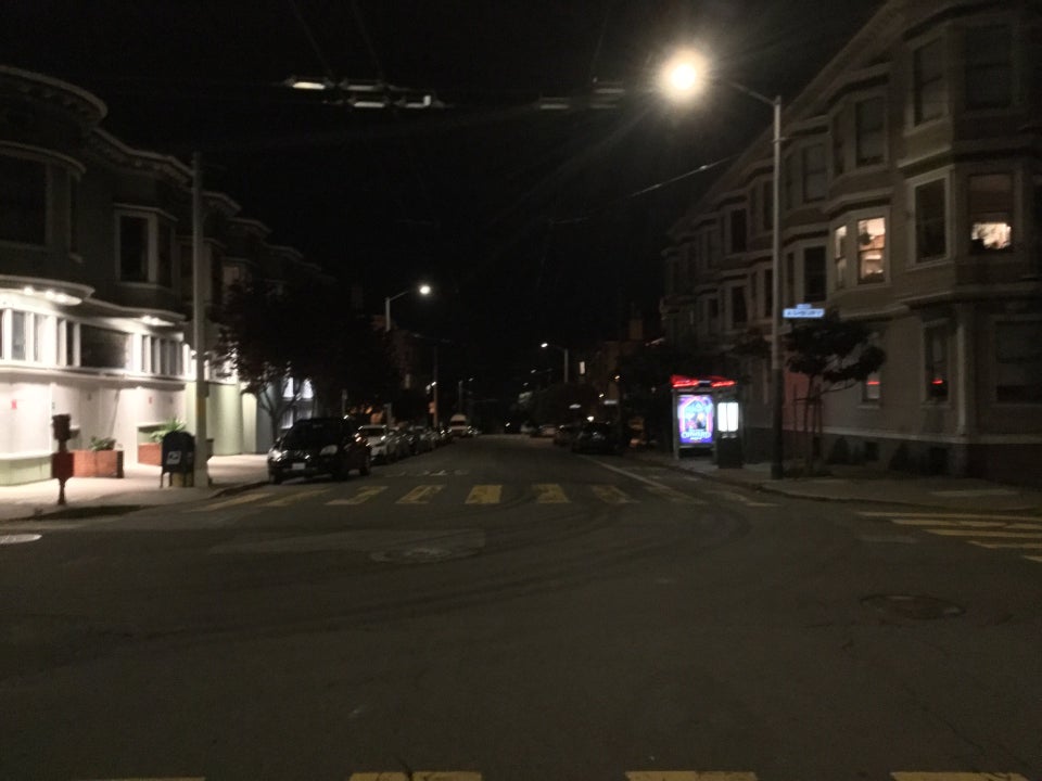 Intersection of Frederick and Ashbury streets at night, looking westward, everything is dark except for streetlamps, lights outside a boarded up Ashbury Market, and a second floor corner apartment on the other side, above a lit bus stop for the number 6 outbound bus.