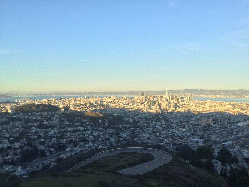 View of sunset-lit downtown San Francisco in the distance as viewed from Twin Peaks, with the hairpin turn below, and immediately below the green hill slope of Twin Peaks summit.