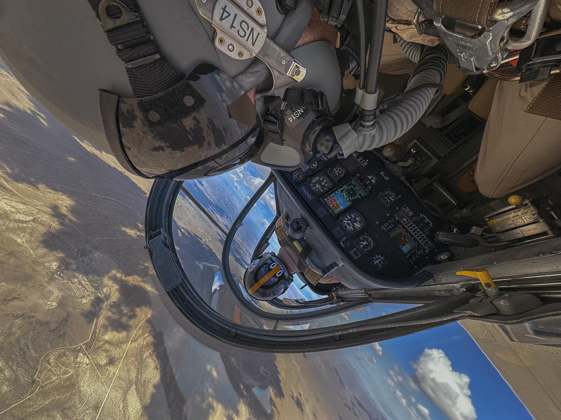 Inside of an aircraft cockpit is shown from the upside down perspective with two men in tan flight suits sitting inside. The side of one helmet, oxygen mask and visor is seen for one of the two men as well as controls inside the aircraft. The second helmet is seen from the back as the man sitting in the front is piloting the aircraft. You can see land below through the window of the aircraft. 