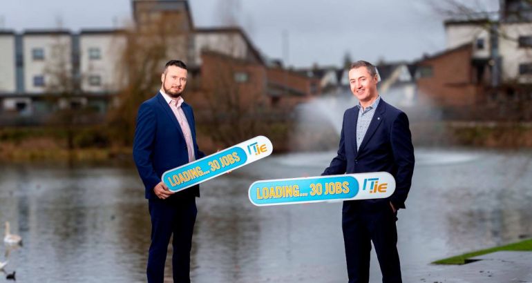 Two men wearing suits stand in front of a canal holding signs advertising 30 new jobs from IT.ie. They are Wayne Morgan and Eamon Gallagher.