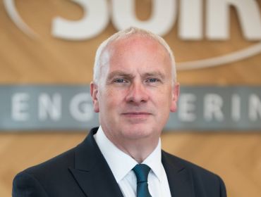 Headshot of Suir Engineering CEO John Kelly wearing a black suit and standing in front of a wall that has the Suir logo on it.