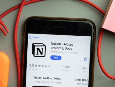 A phone displaying the Notion app download screen. It is lying on a grey table with a red cord and a pink notepad and orange eraser.
