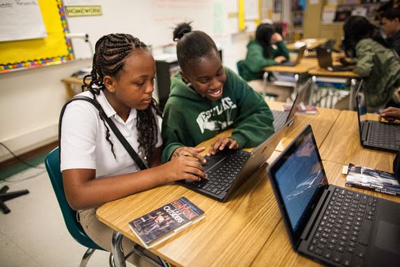 Two secondary school girls share a laptop in a classroom. One girl, wearing a white polo, is focused on the screen. Her friend, in a green hoodie, smiles at the screen.