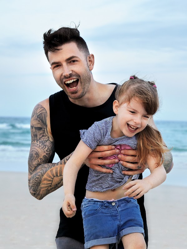 Man in a black vest holding his daughter on the beach and both are smiling