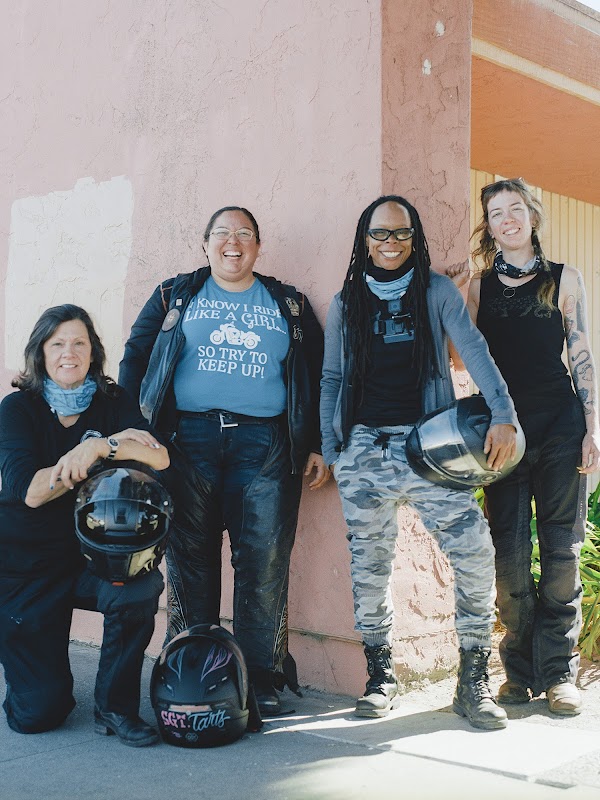 Four women wearing motorcycle gear are standing in a line smiling. Two of them are holding motorcycle helmets.