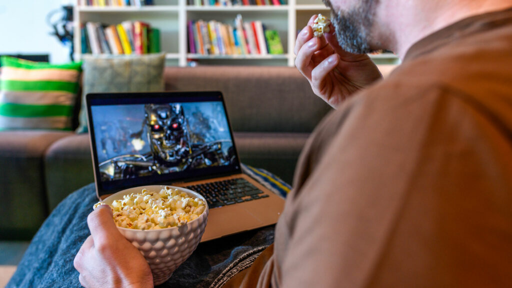 Man sitter vid en laptop och tittar på en ai-genererad bild, han äter popcorn.