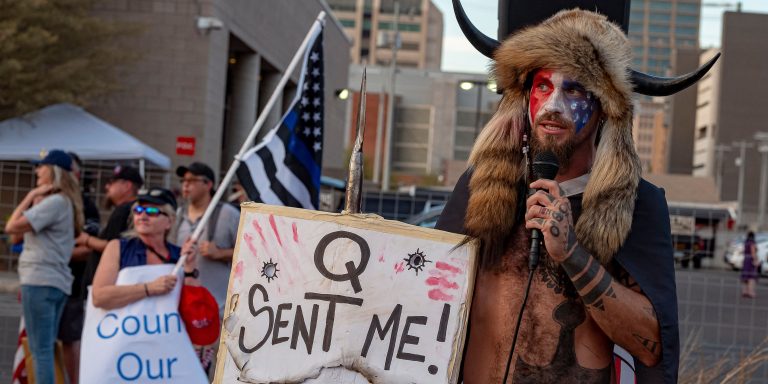 Jake Angeli, known as the "QAnon Shaman,"  holds a sign reading "Q Sent Me" as supporters of US President Donald Trump gather to protest outside the Maricopa County Election Department as counting continues after the US presidential election in Phoenix, Arizona, on November 5, 2020. President Donald Trump erupted on November 5 in a tirade of unsubstantiated claims that he has been cheated out of winning the US election as vote counting across battleground states showed Democrat Joe Biden steadily closing in on victory. (Photo by OLIVIER  TOURON / AFP) (Photo by OLIVIER  TOURON/AFP via Getty Images)