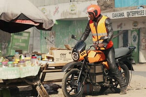 A man with helmet and west standing on an orange electric motorbike on a african market. 